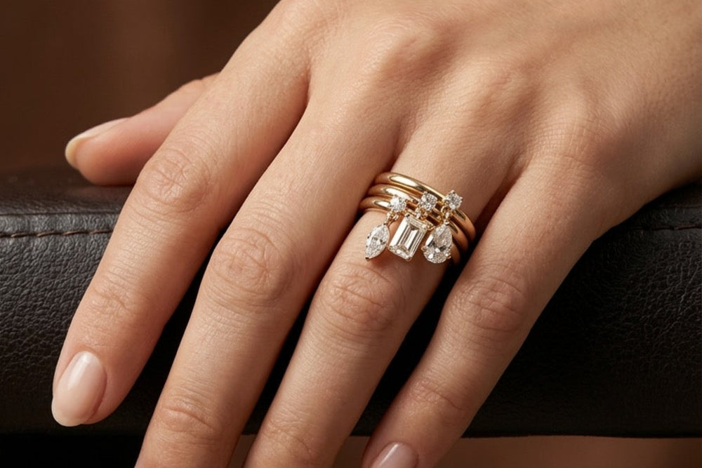 A close-up of a hand wearing a stack of three rings, including a gold band with a diamond cluster setting, against a dark brown background.
